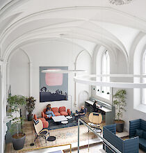 La imagen muestra una habitación alta con techo abovedado y una combinación de elementos de diseño históricos y modernos. A la izquierda de la sala hay grandes plantas que dan a la estancia un ambiente fresco. En el centro hay una acogedora zona de asientos con muebles tapizados en rojo y varias mesas pequeñas. Dos personas están sentadas en el sofá y charlan. Al fondo, una mujer y un hombre están ocupados en un largo escritorio con ordenadores portátiles. A la derecha hay modernos nichos de asientos tapizados. Grandes lámparas circulares cuelgan del techo, iluminando el espacio de manera uniforme. Las paredes son blancas y hay grandes ventanas arqueadas por las que entra mucha luz natural. Una gran obra de arte cuelga de la pared del fondo. El suelo está cubierto por una alfombra decorativa a juego con el moderno mobiliario.