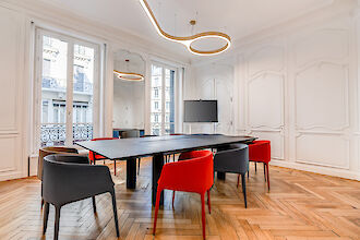 La imagen muestra una moderna sala de reuniones con una gran mesa rectangular negra en el centro. Alrededor de la mesa hay sillas de distintos colores, principalmente rojas y grises. La sala tiene paredes blancas con paneles decorativos y grandes ventanales por los que entra mucha luz natural. Del techo cuelga una llamativa lámpara curva que conecta varias luces en forma de anillo. En un lado de la sala hay una pantalla plana en la pared. El suelo está revestido de parqué en espiga, lo que confiere a la sala un ambiente elegante.