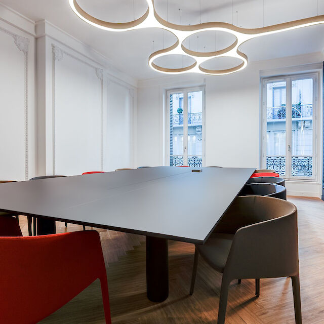 The picture shows a modern conference room with a large, triangular table in the centre. Around the table are several chairs in different colours, including red and grey. The walls of the room are white, with one wall painted in a deep blue colour. An eye-catching, modern light with several round, interconnected rings hangs from the ceiling, illuminating the room. Two large windows let in natural light and offer a view of the building opposite. The floor is covered with wooden parquet, which gives the room a warm atmosphere.