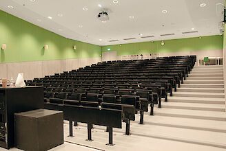 La imagen muestra una moderna sala de conferencias con filas de butacas equipadas con asientos negros plegables. Las paredes son de color verde claro y la mitad inferior está revestida de paneles de madera gris claro. En el techo se ven numerosos focos redondos empotrados. En la parte delantera izquierda de la imagen hay un atril. Al final de la sala hay dos puertas dobles, y en la pared cuelga una señal de salida de emergencia que apunta en la dirección de las puertas. La sala parece limpia y ordenada, con una estética clara y funcional.