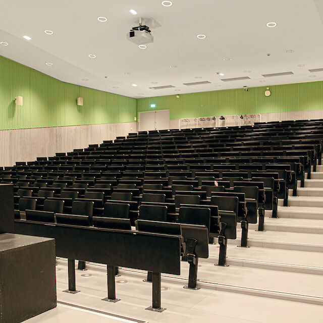 La imagen muestra una moderna sala de conferencias con filas de butacas equipadas con asientos negros plegables. Las paredes son de color verde claro y la mitad inferior está revestida de paneles de madera gris claro. En el techo se ven numerosos focos redondos empotrados. En la parte delantera izquierda de la imagen hay un atril. Al final de la sala hay dos puertas dobles, y en la pared cuelga una señal de salida de emergencia que apunta en la dirección de las puertas. La sala parece limpia y ordenada, con una estética clara y funcional.