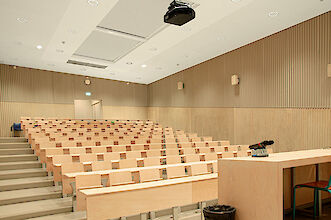 La imagen muestra un auditorio con filas escalonadas de asientos de madera clara. Los asientos tienen un diseño clásico con mesas integradas. En la parte delantera de la sala hay un atril con un micrófono y una pequeña consola encima. Las paredes son de color beige neutro con paneles verticales de madera que dan estructura a la sala. En el techo hay focos empotrados redondos y compactos y un proyector. Al fondo se ve una puerta cerrada sobre la que cuelga una señal luminosa de salida de emergencia. La sala parece tranquila y funcional.