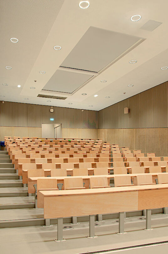 Hörsaal mit Holzverkleidung, hellen Sitzreihen und ringförmigen Ringo Star Einbauleuchten.
