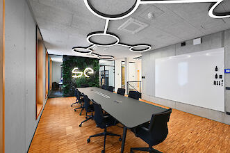 Das Bild zeigt einen modernen Konferenzraum mit einem langen, rechteckigen Tisch in der Mitte, der von schwarzen Bürostühlen umgeben ist. Die Wände und die Decke sind in einem minimalistischen, industriellen Stil gehalten, mit Betonoberflächen und integrierten Lichtquellen. An der Decke befinden sich auffällige, kreisförmige LED-Leuchten, die in einem verbundenen Muster angeordnet sind. Auf der rechten Seite des Raumes befindet sich eine große weiße Schreibtafel, die für Präsentationen oder Notizen genutzt werden kann.

Am Ende des Raumes ist eine auffällige Wand mit vertikalen Pflanzen bedeckt, die eine lebendige grüne Fläche schafft. Inmitten dieser Pflanzenwand leuchtet ein neonartiges „SE“-Logo in Weiß, was dem Raum ein modernes und stilvolles Ambiente verleiht. Links im Bild gibt es große Fenster mit Holzrahmen, die viel natürliches Licht hereinlassen und den Raum mit einer warmen Atmosphäre erfüllen. Der Boden besteht aus hellem Holzparkett, das den industriellen Charakter des Raumes ergänzt und gleichzeitig Wärme und Komfort ausstrahlt.