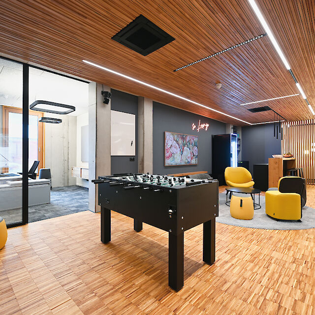 The picture shows a modern lounge and break area in an office with a warm, inviting atmosphere. In the foreground is a black foosball table on a floor of light-coloured wooden parquet. The ceiling is clad with horizontal wooden slats with linear LED lights running through them. To the right of the picture is a cosy seating area with yellow and black armchairs and a round grey carpet. On the wall behind the seating area hangs a bright neon sign with the words ‘Lounge’ and a picture with floral motifs. On the left, two workstations with desks and chairs are visible through a glass wall, which are located in a separate room. The rear wall of this room is in a raw concrete style, which contrasts with the warm wooden elements. The entire room exudes a modern, stylish yet relaxed atmosphere.