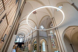 La imagen muestra el interior de una iglesia moderna remodelada con altos techos abovedados y arcos góticos. Grandes luces LED en forma de anillo cuelgan del techo e iluminan suavemente la sala. Al fondo se ven vidrieras llenas de color y detalles, complementadas por la moderna iluminación. En los laterales de la sala hay paredes de madera clara y barandillas de cristal, que aportan un acento moderno. La arquitectura conserva la estructura tradicional de la iglesia, mientras que la iluminación y el diseño dan a la sala un aire contemporáneo.