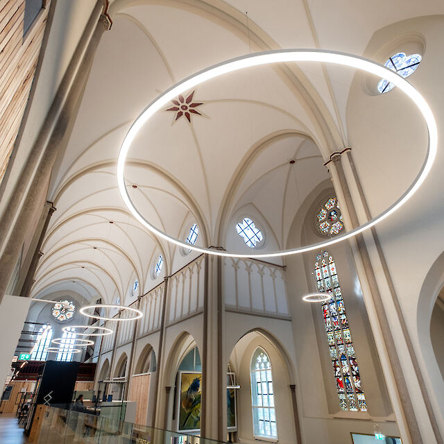 The picture shows the interior of a modern remodelled church with high vaulted ceilings and Gothic arches. Large, ring-shaped LED lights hang from the ceiling and gently illuminate the room. Colourful, detailed stained glass windows can be seen in the background, complemented by the modern lighting. On the sides of the room are walls made of light-coloured wood and glass railings, which provide a modern accent. The architecture preserves the traditional structure of the church, while the lighting and design give the room a contemporary feel.