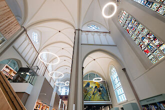 La imagen muestra el interior de una iglesia moderna remodelada con altos techos abovedados y arcos góticos. Grandes luces LED en forma de anillo cuelgan del techo e iluminan suavemente la sala. Al fondo se ven vidrieras llenas de color y detalles, complementadas por la moderna iluminación. En los laterales de la sala hay paredes de madera clara y barandillas de cristal, que aportan un acento moderno. La arquitectura conserva la estructura tradicional de la iglesia, mientras que la iluminación y el diseño dan a la sala un aire contemporáneo.