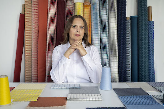 La diseñadora textil Aleksandra Gaca se sienta a una mesa con muestras de telas, rodeada de coloridos rollos de tejidos acústicos.