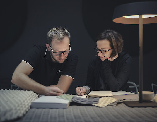 Dos diseñadores examinan muestras de tejido para la luminaria Serenity bajo una luminaria de sobremesa Beam Me Up.
