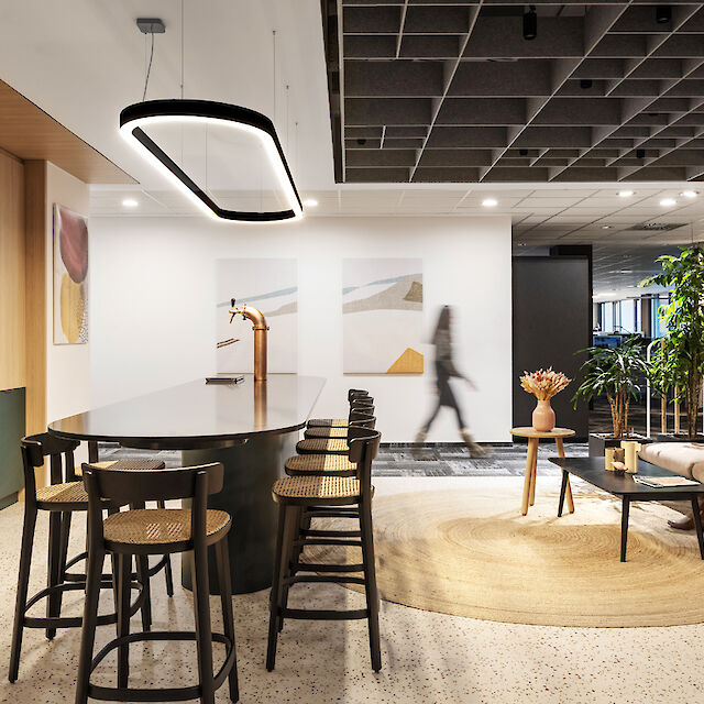 The picture shows a modern, stylish office space with a cosy atmosphere. In the foreground is a long, oval table island with several black bar stools and a golden beer dispenser. To the left is a green kitchen unit with a fully automatic coffee machine. A person walks blurred in the background, while another person sits relaxed on a beige-coloured sofa on the right, working on a laptop. The room is brightly lit by an eye-catching oval pendant light above the table. The ceiling consists of a grey, grid-like acoustic system and the floor has a light, dotted texture.